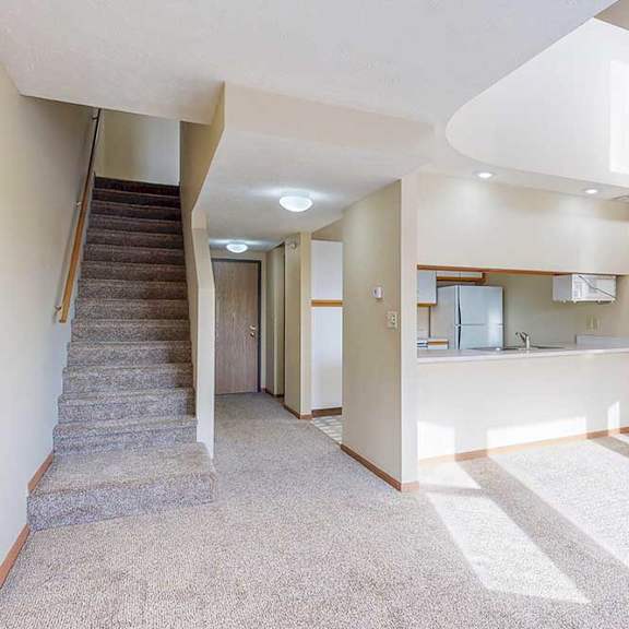 a carpeted living room with access to the kitchen via a pass-through window countertop, and a staircase to the upper loft area.