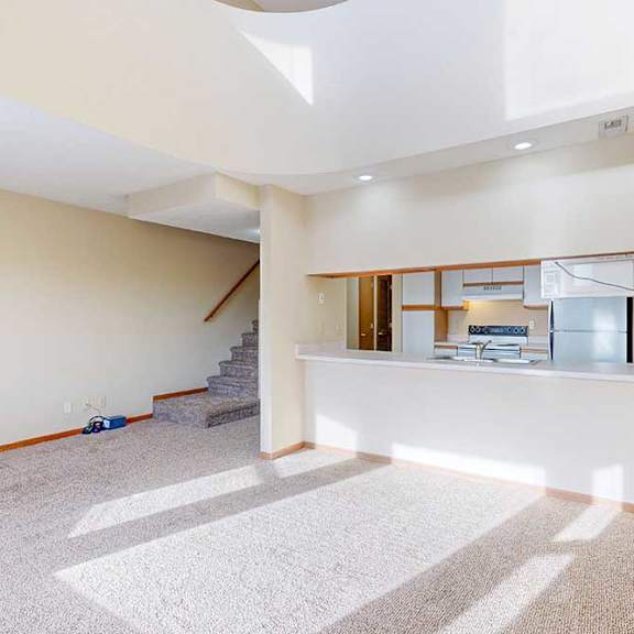 a carpeted living room with access to the kitchen via a pass-through window countertop, and a staircase to the upper loft area.