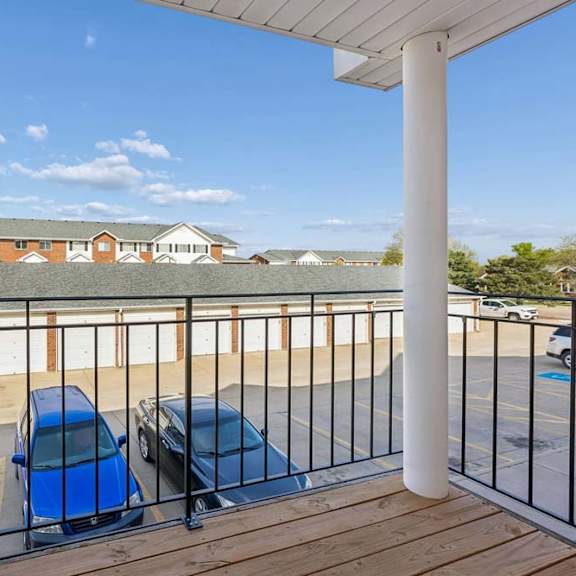 Thumbnail 16 of 16 Covered apartment balcony with wood decking, white support columns, metal railing, and views of the community parking area and neighboring apartment buildings under a clear sky.