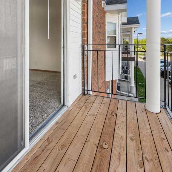 Thumbnail 15 of 16 Covered apartment balcony with wood decking, a sliding glass door, brick accents, white support columns, and views of the surrounding apartment community.