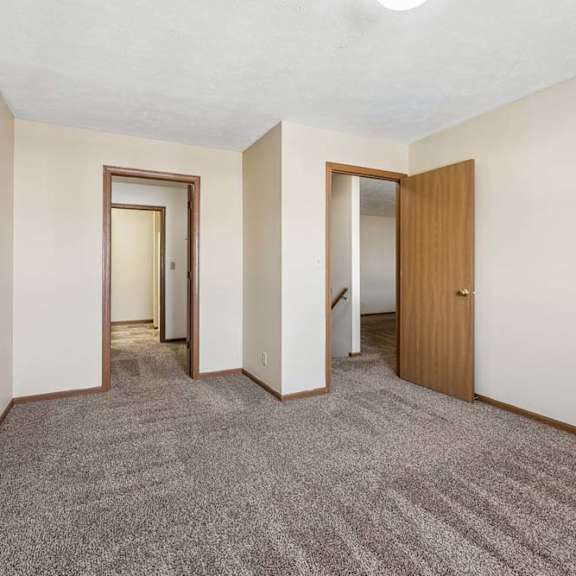 Thumbnail 9 of 16 Spacious apartment bedroom with neutral carpet, beige walls, wood trim, and open doorways leading to the hallway, closet area, and staircase.