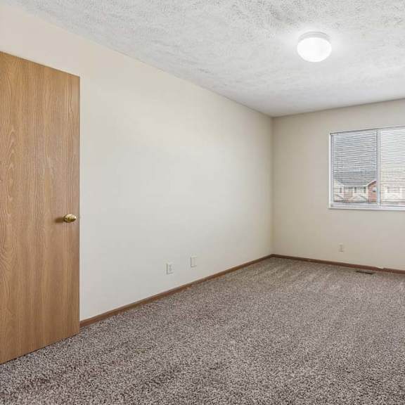 Thumbnail 13 of 16 Bright, empty apartment bedroom with neutral carpet, beige walls, wood trim, and a window with blinds providing natural light.