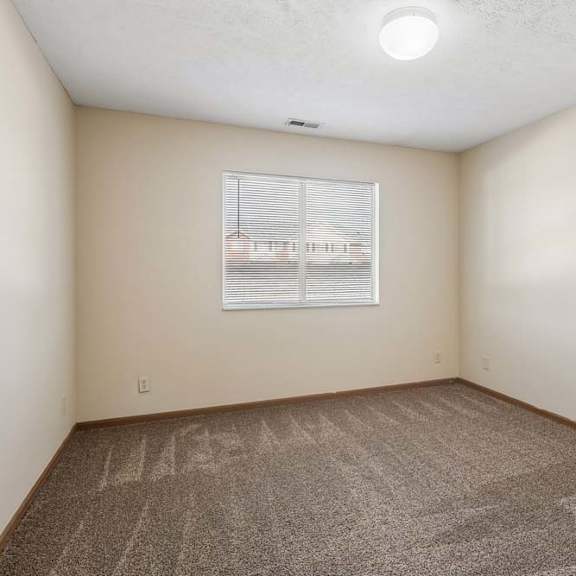 Thumbnail 8 of 16 Bright, empty apartment bedroom with neutral carpet, beige walls, wood trim, and a window with blinds that brings in natural light.
