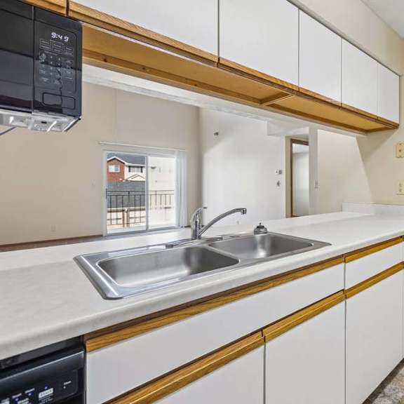 Thumbnail 3 of 16 Open apartment kitchen with white cabinets, a double-basin sink, laminate countertops, and a pass-through breakfast bar overlooking the adjacent living area and staircase.