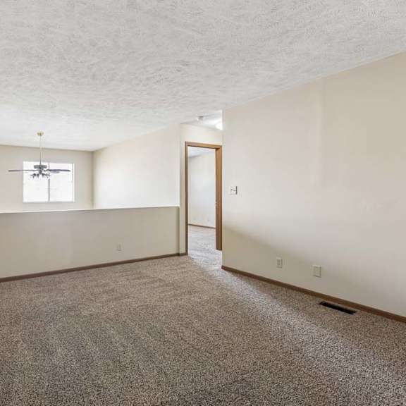 Thumbnail 6 of 16 Spacious loft area in an apartment with neutral carpet, a half wall overlooking the lower level, a window for natural light, and an open doorway leading to the hallway.