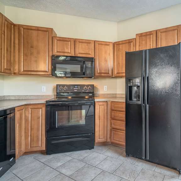 Thumbnail 7 of 16 A black refrigerator stands in a kitchen with wooden cabinets and a black microwave.