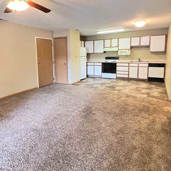 Thumbnail 4 of 11 a spacious living room with a carpeted floor and a ceiling fan, and a kitchen with white and brown cabinets in the background