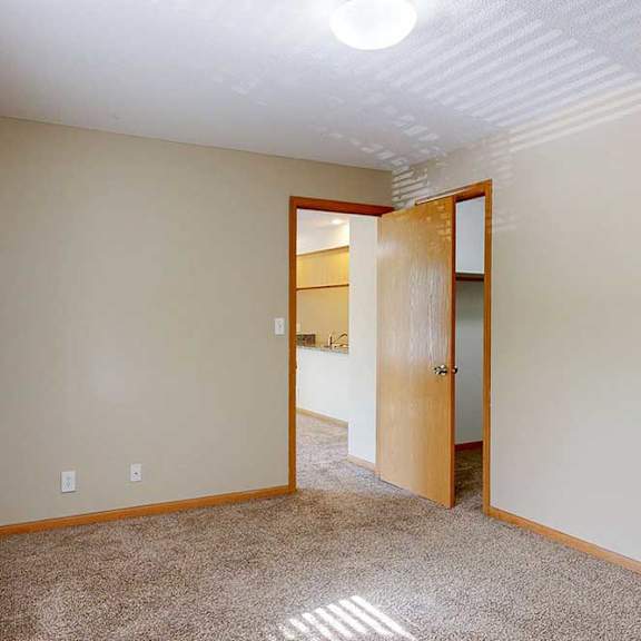 Thumbnail 12 of 22 Carpeted apartment bedroom with neutral paint, ceiling light fixture, wood-trimmed door, and view into adjoining living area.