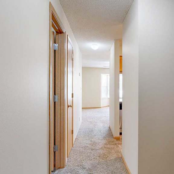 Thumbnail 2 of 22 Apartment hallway with beige walls, wood-trimmed doors, carpeted flooring, and overhead lighting leading toward the living area.
