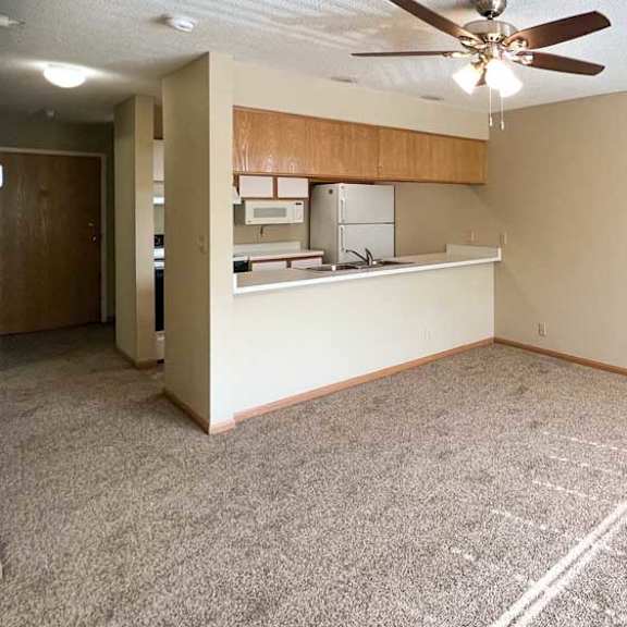 Thumbnail 4 of 9 a carpeted living room with a ceiling fan, and a kitchen with a windowed countertop for dining seating