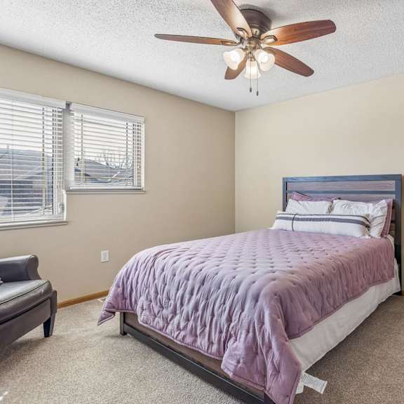 Thumbnail 20 of 29 Bright apartment bedroom with neutral carpet, large corner windows, ceiling fan, and a queen-size bed, offering a comfortable and airy interior.