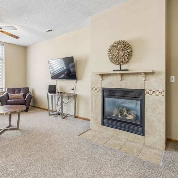 Thumbnail 4 of 29 Cozy apartment living room with neutral carpet, tiled fireplace, large window with blinds, ceiling fan, and seating area with a wall-mounted TV.