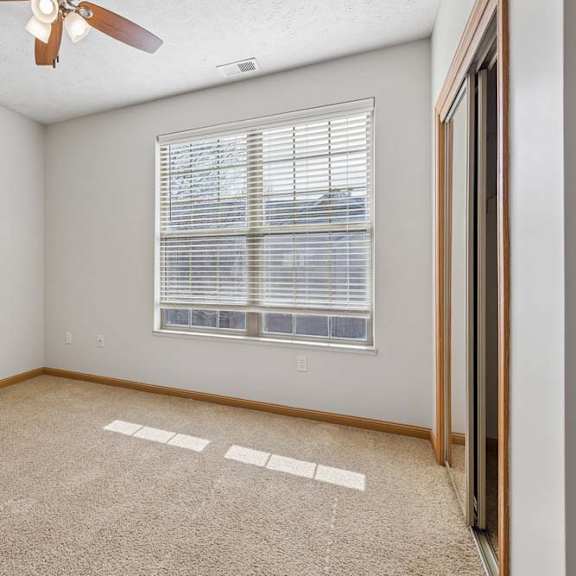Thumbnail 18 of 19 Sunlit apartment bedroom with a wide window, mirrored sliding closet doors, neutral carpeting, and a ceiling fan.