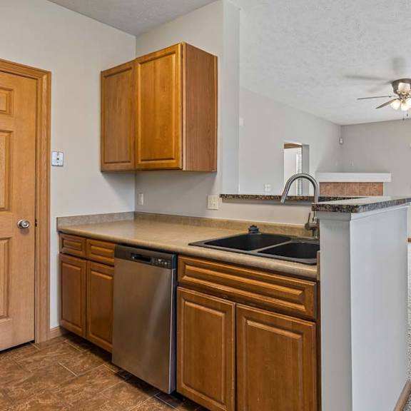 Thumbnail 15 of 19 Open-concept apartment kitchen and living area with wood cabinetry, breakfast bar, dishwasher, ceiling fan, and sliding glass door.