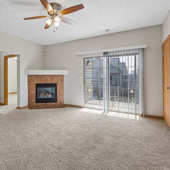 Thumbnail 16 of 19 Bright, empty apartment living room with beige carpet, tiled fireplace, ceiling fan, and sliding glass doors opening to a balcony.