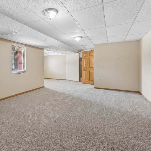 Thumbnail 14 of 17 Spacious, empty lower-level room with neutral carpet, drop ceiling, recessed lighting, windows with blinds, and white brick wall.
