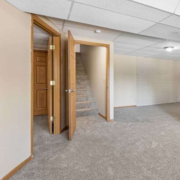 Thumbnail 16 of 17 Empty lower-level room with neutral carpet, drop ceiling, white brick walls, overhead lighting, and stair access.