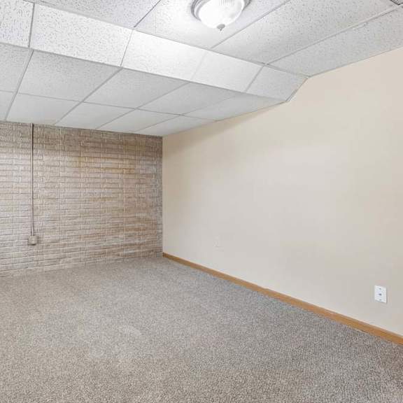 Thumbnail 15 of 17 Empty lower-level room with neutral carpet, exposed brick accent wall, drop ceiling, and overhead lighting.