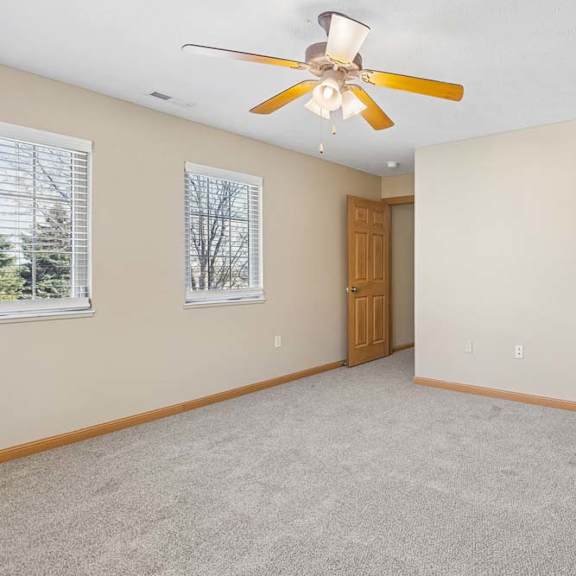 Thumbnail 11 of 17 Bright, empty bedroom with neutral carpet, two windows with blinds, ceiling fan, wood trim, and direct bathroom access.