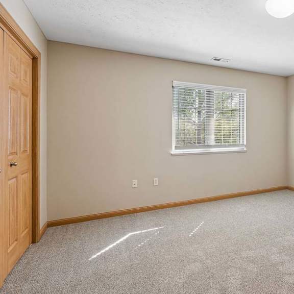 Thumbnail 9 of 17 Bright, empty bedroom with neutral carpet, a large window with blinds, wood trim, and double closet doors.