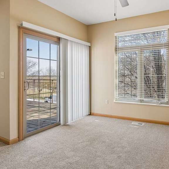 Thumbnail 7 of 17 Bright, empty living area with neutral carpet, large window with blinds, ceiling fan, and sliding glass door to the deck.