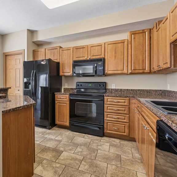 Thumbnail 2 of 17 Kitchen with wood cabinetry, speckled countertops, black appliances, breakfast bar, tile flooring, and a large window.