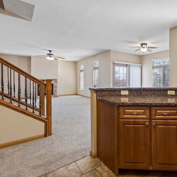 Thumbnail 3 of 17 Open kitchen and living area with wood cabinetry, speckled breakfast bar, tile and carpet flooring, large windows, and staircase.