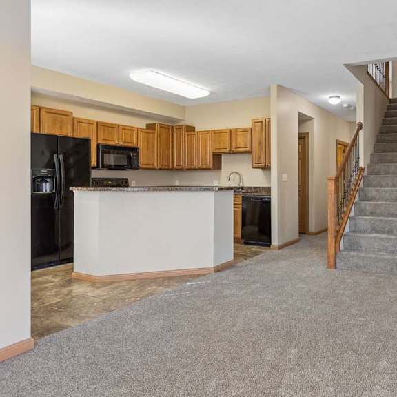 Thumbnail 6 of 17 Open living area with neutral carpet, central staircase, kitchen with wood cabinetry, breakfast bar, and black appliances.
