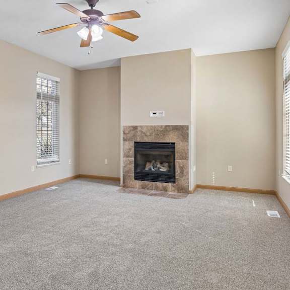 Thumbnail 4 of 17 Bright, empty living room with neutral carpet, tiled fireplace, ceiling fan, and tall windows with blinds.