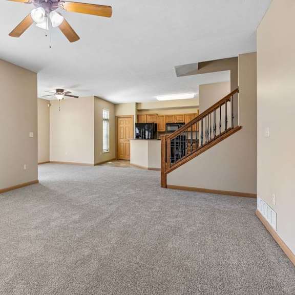 Thumbnail 5 of 17 Spacious, empty living area with neutral carpet, ceiling fans, tall windows, open kitchen, and wood-railed staircase.
