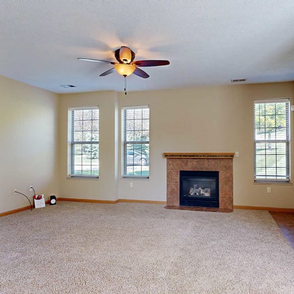 Thumbnail 4 of 28 a living room with a ceiling fan, a brick/tile fireplace, three tall windows, and a tiled entryway.