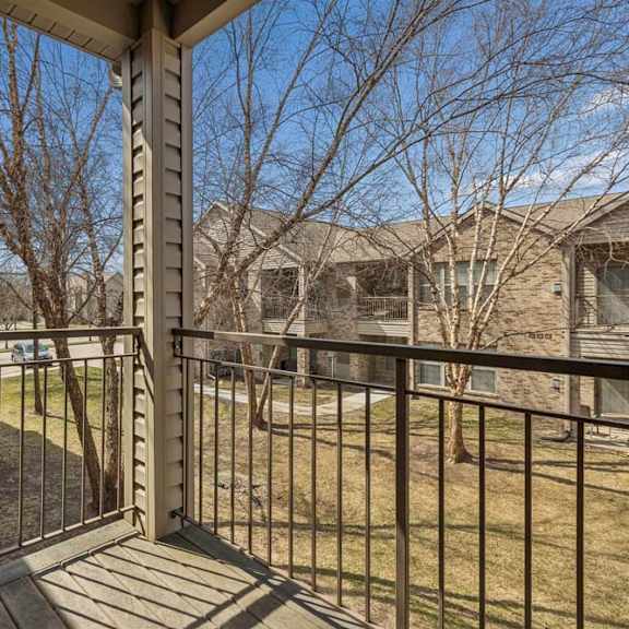 Thumbnail 31 of 31 Covered apartment balcony with metal railing overlooking mature trees, landscaped grounds, and neighboring community buildings.