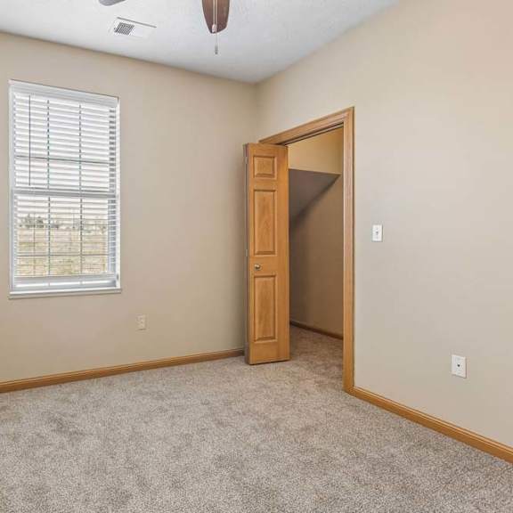 Thumbnail 28 of 31 Cozy apartment bedroom with neutral carpeting, a tall window with blinds, ceiling fan, and wood-trimmed doorway.