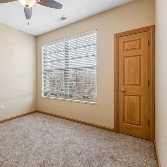 Thumbnail 29 of 31 Bright apartment bedroom with a wide window and blinds, neutral carpeting, ceiling fan, and a wood closet door.