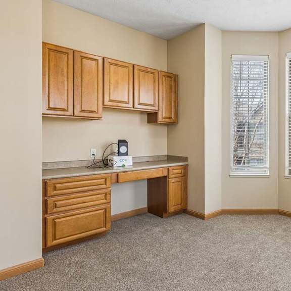 Thumbnail 26 of 31 Bright apartment office nook with built-in wood cabinetry, a desk area, carpeted flooring, and three large windows with blinds.