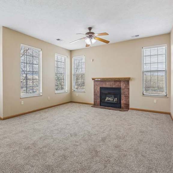 Thumbnail 25 of 31 Bright apartment living room with a corner fireplace, carpeted flooring, ceiling fan, and six tall windows with blinds.