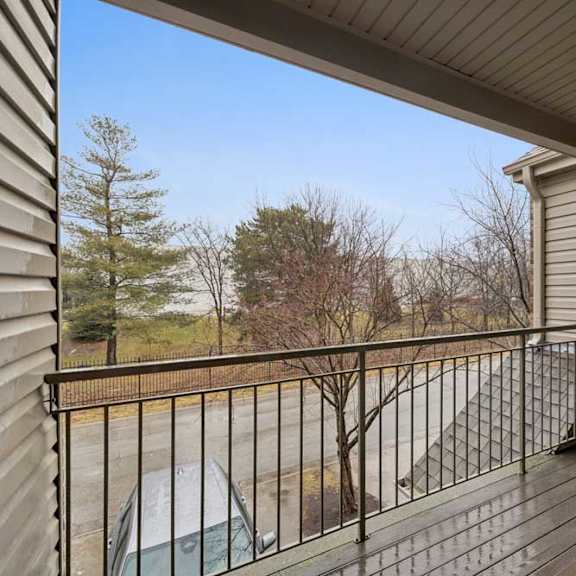 Thumbnail 6 of 19 Covered apartment balcony with wood-look decking and metal railing, overlooking a parking area and mature trees.
