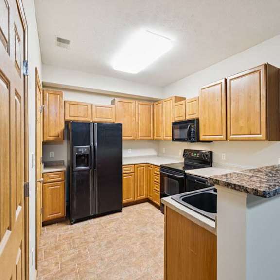 Thumbnail 10 of 19 Apartment kitchen with wood cabinetry, black refrigerator, oven, microwave, and breakfast bar with raised counter.