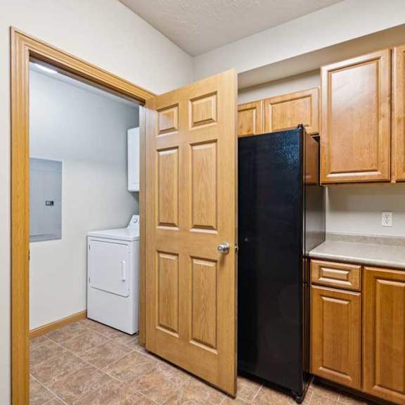 Thumbnail 12 of 19 Apartment kitchen with wood cabinetry and laminate countertops, featuring a black refrigerator and adjacent in-unit laundry closet with washer and dryer.