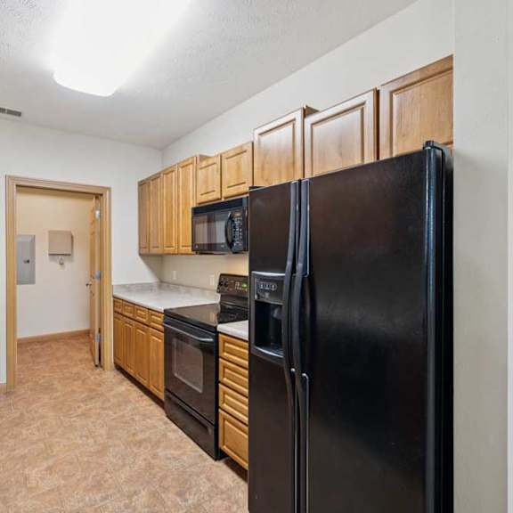 Thumbnail 6 of 19 Galley-style kitchen with natural wood cabinets, black refrigerator, oven and microwave, laminate countertops, and tile flooring.