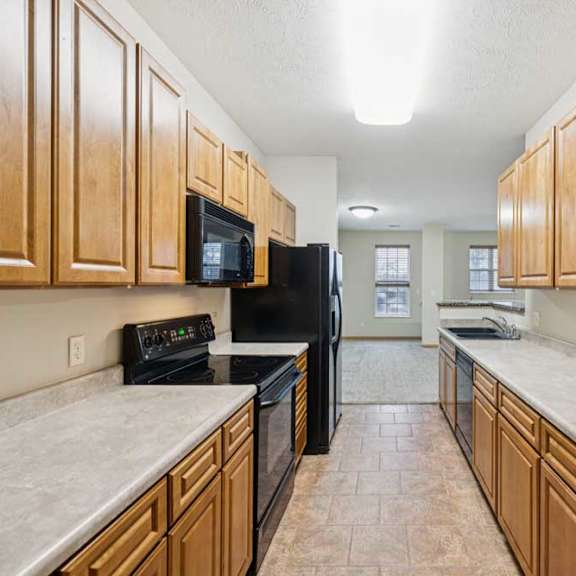 Thumbnail 8 of 19 Galley-style kitchen with natural wood cabinets, laminate countertops, black oven and microwave, and tile flooring leading to open living area.