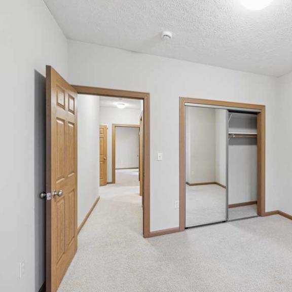 Thumbnail 19 of 20 Bedroom with mirrored closet doors, neutral carpet, and hallway access in a well-lit apartment space.