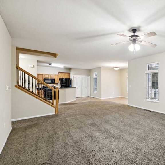 Thumbnail 5 of 25 Spacious open living area with carpet flooring, staircase with wood railing, and kitchen in background.