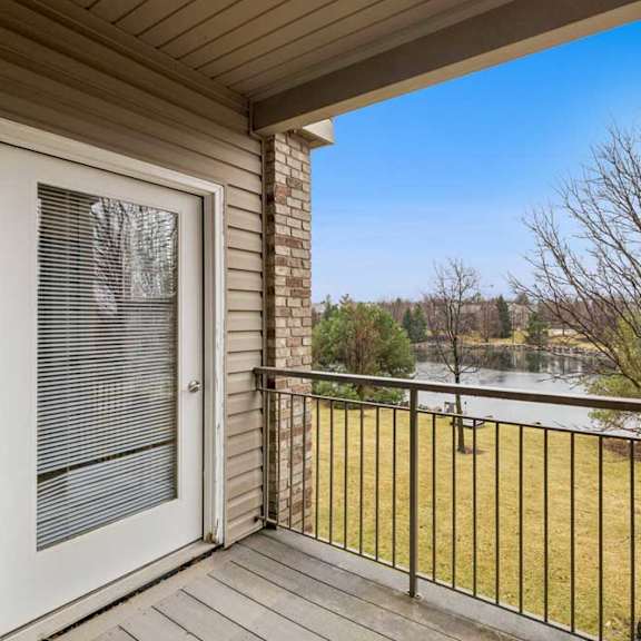 Thumbnail 13 of 21 Private apartment balcony with wood decking and metal railing overlooking a pond and landscaped green space, accessed by a glass patio door.