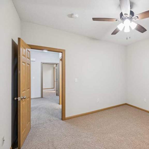 Thumbnail 19 of 21 Empty apartment bedroom with neutral carpet, white walls, wood trim, and ceiling fan, connected to an adjoining hallway.