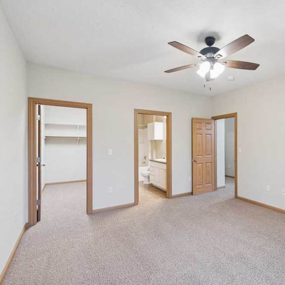 Thumbnail 6 of 21 Spacious bedroom with neutral carpet, ceiling fan, and wood trim, featuring an en-suite bathroom and large walk-in closet through separate doorways.