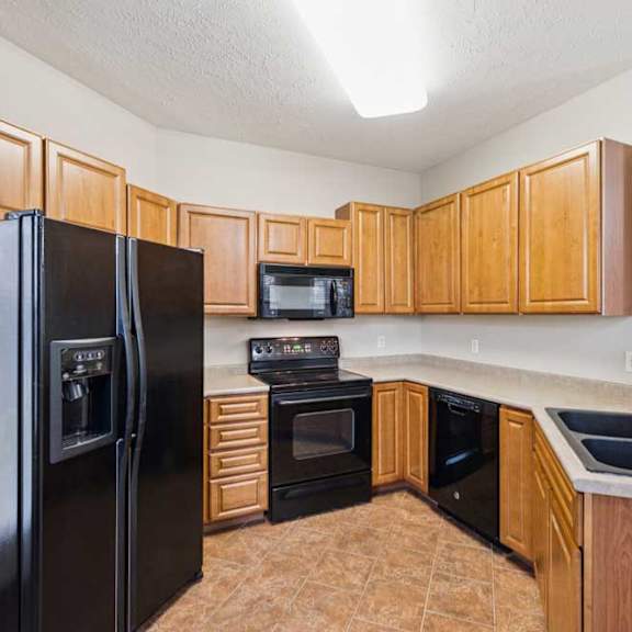 Thumbnail 14 of 21 U-shaped apartment kitchen with wood cabinetry, black refrigerator, oven and microwave, laminate countertops, and tile flooring.