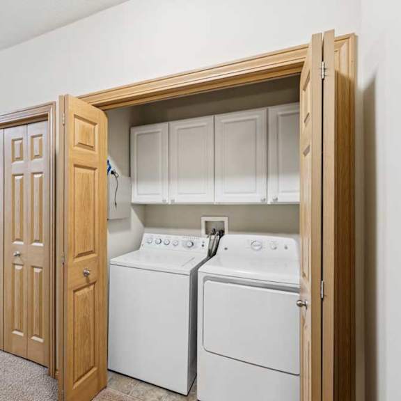 Thumbnail 3 of 21 In-home laundry closet with side-by-side washer and dryer, upper storage cabinets, tile flooring, and wood bi-fold doors in a carpeted hallway.