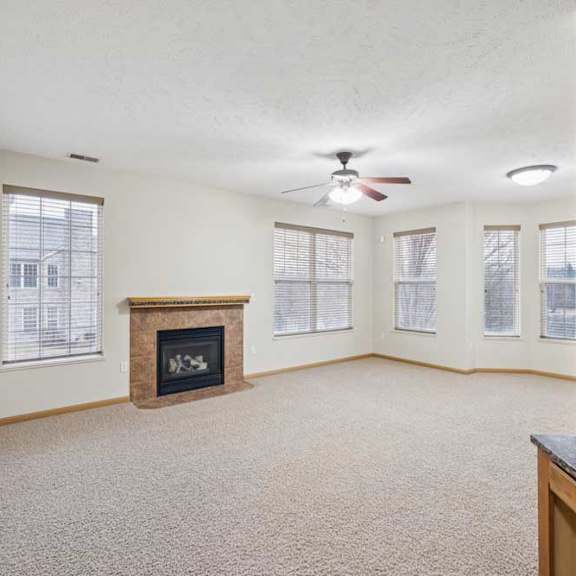 Thumbnail 9 of 21 Open living room interior with neutral carpet, multiple windows, ceiling fan, and a corner fireplace with tile surround and wood mantel.