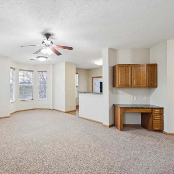 Thumbnail 10 of 21 Open apartment living and dining area with neutral carpet, multiple windows, ceiling fan, and built-in desk with upper cabinets near the kitchen.